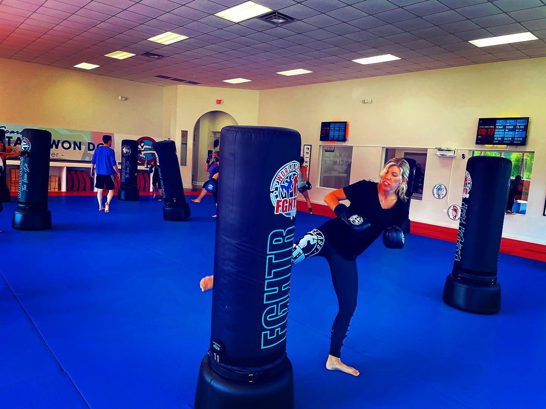 Woman kicking a heavy bag during a FGHTR Fitness Kickboxing class with an Impact Wrap leaderboard in background.